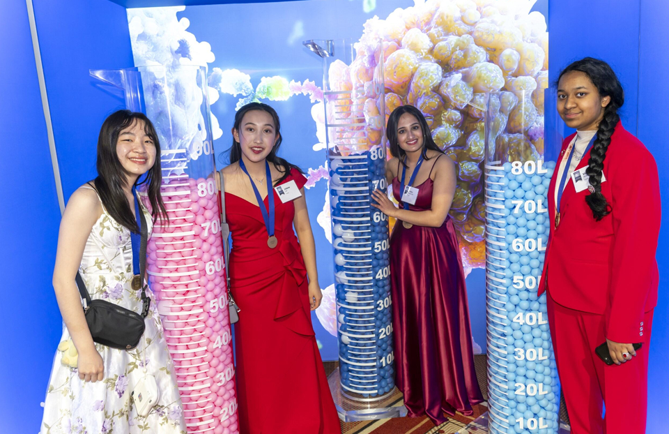 Regeneron STS 2025 finalists Melody Hong, Jolene Cao, Prisha Bhat, and Vidya Ambati standing beside large graduated cylinders at the STS gala.
