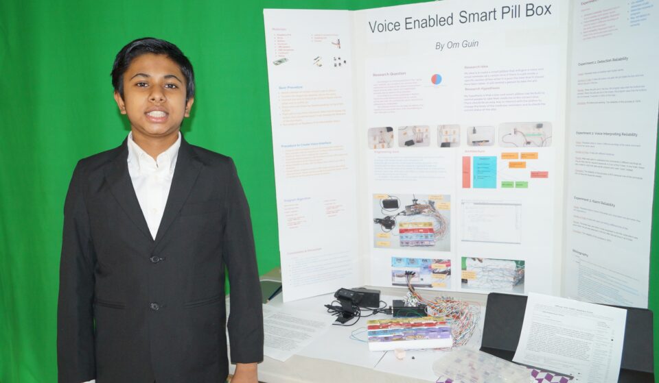 Om Guin stands in front of his project, a voice-enabled smart pillbox aimed to remind seniors to take their medication