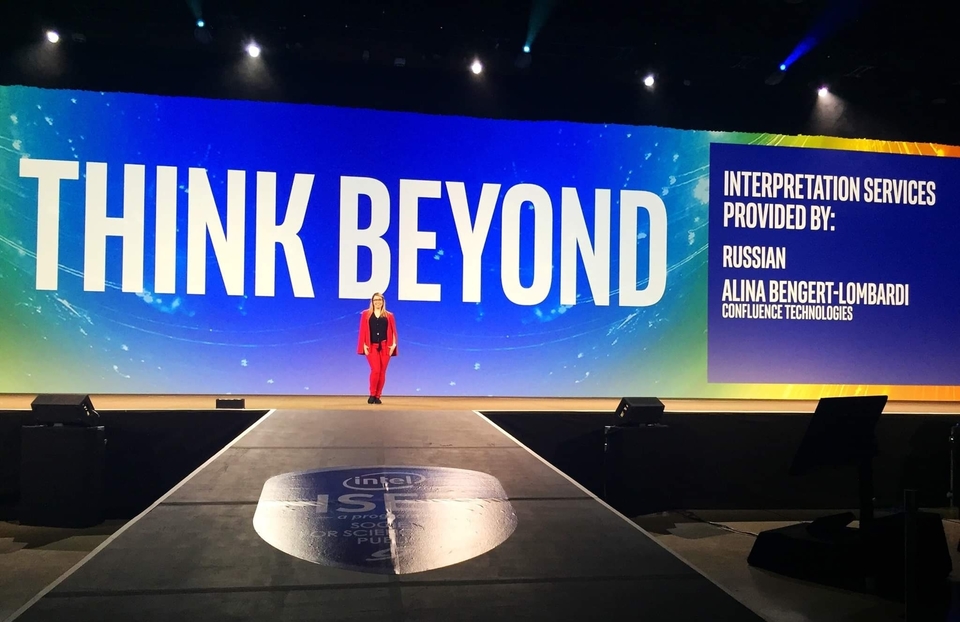Alina Bengert-Lombardi stands on stage at ISEF. She is in front of a large screen that identifies her as providing Russian interpretation services.