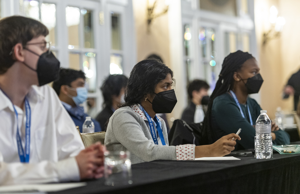 Three Regeneron Science Talent Search finalists, wearing masks, sit at a table facing a speaker (out of frame) during STS finals week