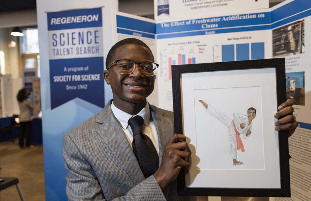 Elijah Burks, an STS 2022 alumni, poses with a hand drawn portrait of himself in front of his project board at Public Day 2022.