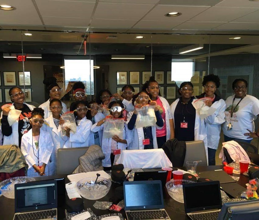 A group of young girls gather for a picture wearing white lab coats and goggles.