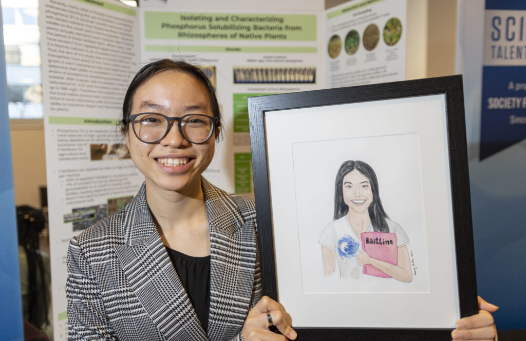 Krystal Li, a 2022 STS alumni, poses with a hand drawn portrait of herself in front of her project board at Public Day 2022.