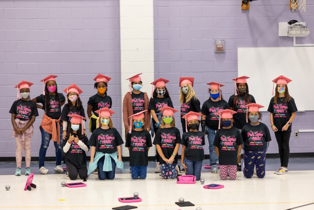 A group of young girls pose for a picture in a gymnasium, wearing pink graduation caps.