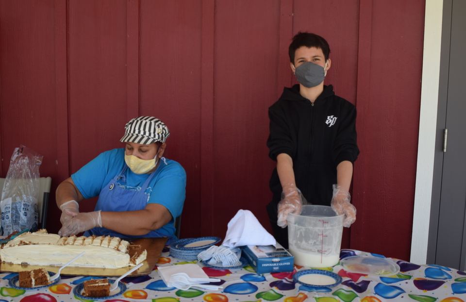 Two participants behind a table, one sitting and cutting a cake, the other standing with a bucket of milk or cream, at Rural Resources Farm & Food Education Center