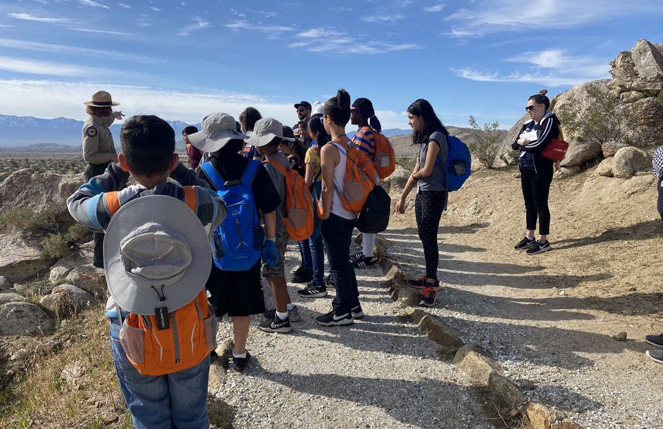 Standing in a desert landscape, a group of about 15 students listen to a park ranger who is pointing toward the horizon