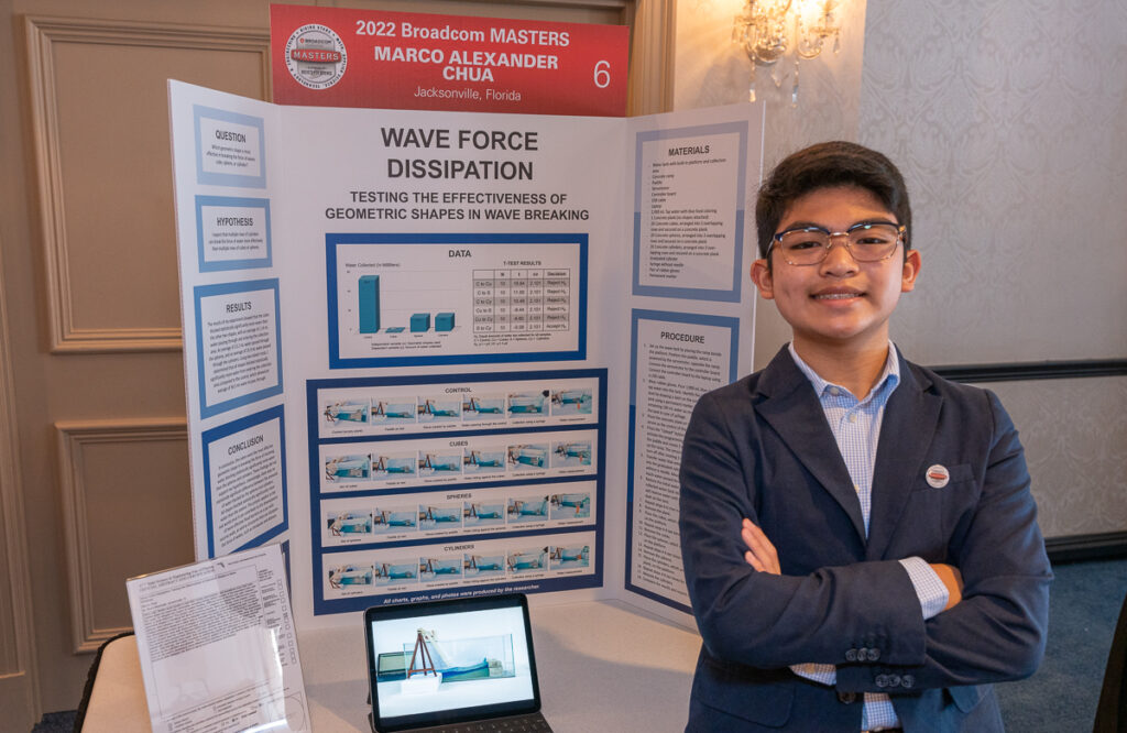 2022 Broadcom MASTERS Finalist Marco Chua smiles in front of his project board, displaying his research on Wave Force Dissipation: Testing the effectiveness of geometric shapes in wave breaking
