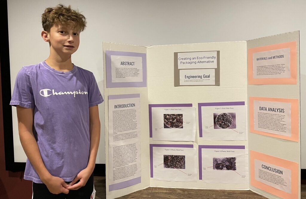 Student Finnegan Miller stands next to his project board, which displays his research on using mushrooms as an eco-friendly packaging option. He won the Lemelson Early Inventor Prize for his research.