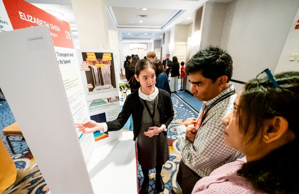 Elizabeth Shen stands at her project poster, speaking with two adults about her work.