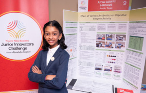 Thermo Fisher JIC finalist crosses arms in front of poster board. Finalist is wearing a blue blazer suit with a Thermo Fisher JIC lapel pin.