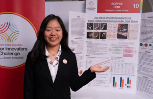 Thermo Fisher JIC finalist poses in front of their poster board. Finalist is wearing a black blazer with a Thermo Fisher JIC lapel pin.