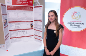 Thermo Fisher JIC finalist poses in front of poster board. Finalist is wearing a black dress with a Thermo Fisher JIC lapel pin.