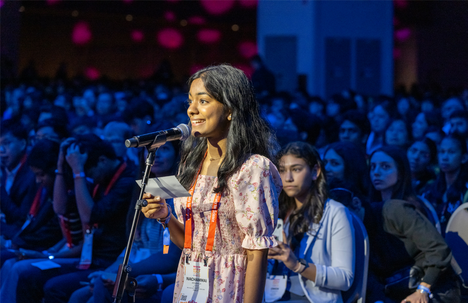 A girl wearing a dress and dark brown hair is holding up a question card at a microphone to ask panelists a question