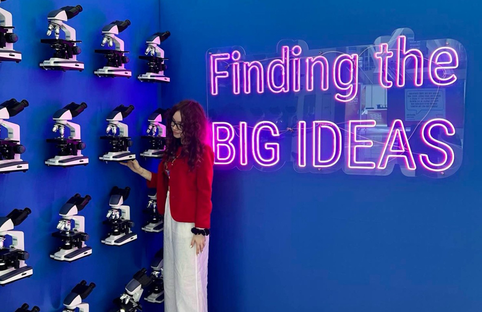 On the left of the image is a wall of microscopes and a girl in a red blazer looking at one. On the right of the image is neon lettering that reads "Finding the big ideas"
