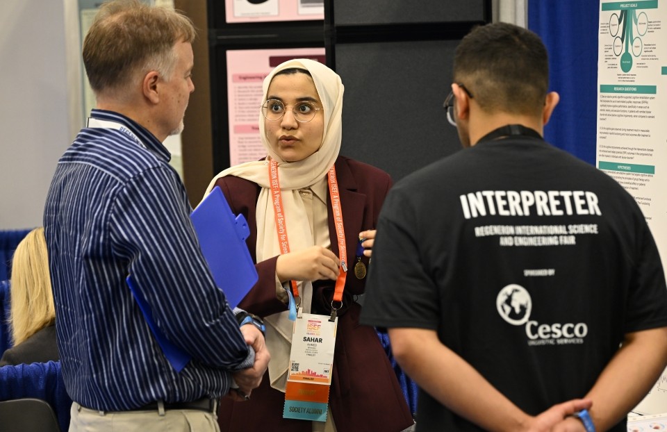 An interpreter helps a Regeneron ISEF finalist present her work to a judge in front of her project poster.