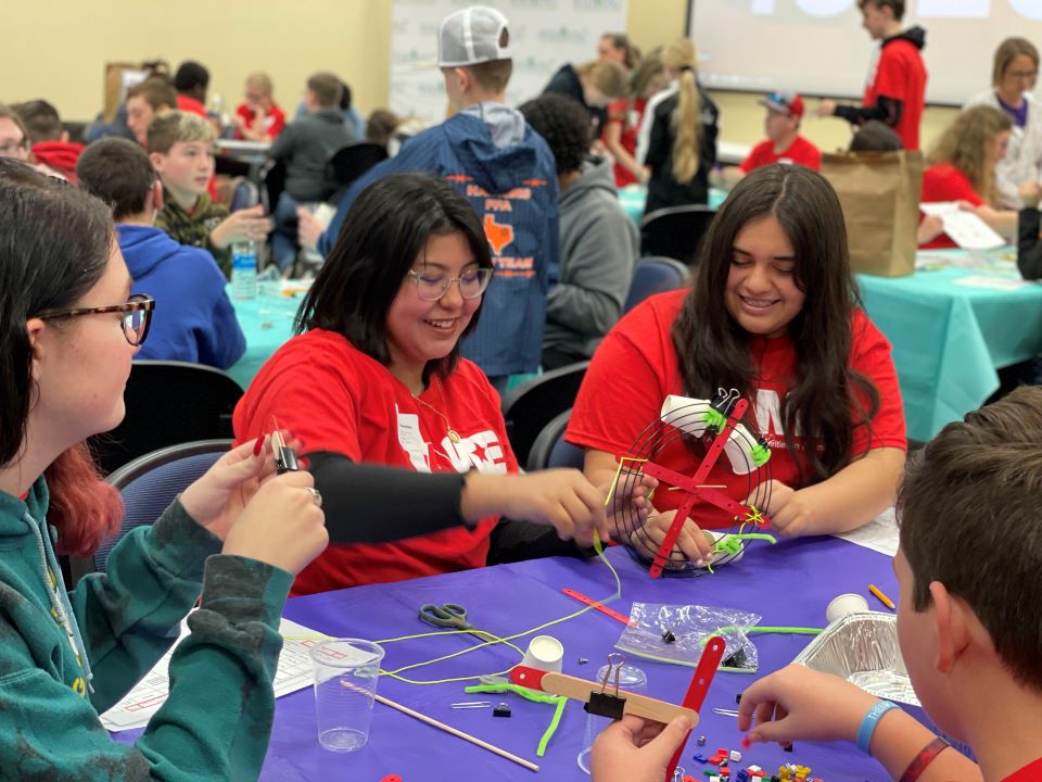 Two girls girls in red shirts smile while working on an engineering project.