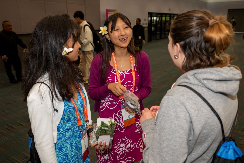 2024 ISEF - Finalists with flowers behind their ears smile while exchanging pins with another student.