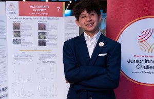 Thermo Fisher JIC finalist crosses arms in front of poster board. Finalist is wearing a black suit with a Thermo Fisher JIC lapel pin. 