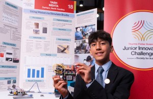 Thermo Fisher JIC finalist holds braille device in front of poster board. Finalist is wearing a black suit with a blue tie and a Thermo Fisher JIC lapel pin. 