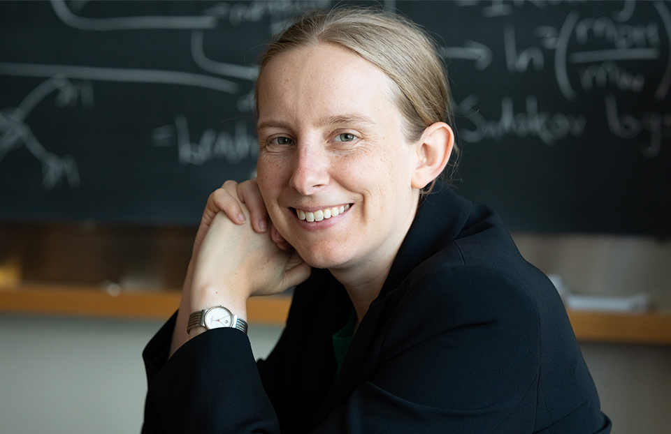 Tracy Slatyer is staring at the camera, smiling. There is a black board with equations written on in the background. Slatyer is wearing a black jacket and silver watch. Her light brown (or dark blonde) hair is pulled back in a ponytail. Physicist Tracy Slatyer imagines different ways dark matter might interact with ordinary matter, helping researchers learn how to look for it.