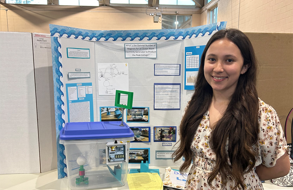 A girl with long brown hair poses with a smile in front of her project board.