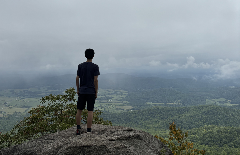 2025 STS Finalist Ray Zhang looking over a cliff while standing on a rock. There is a grey cloudy sky that lays above green forestry trees.
