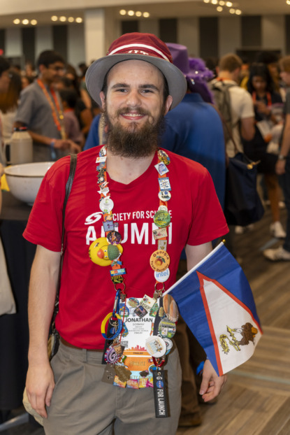 ISEF finalist posing and wearing a lanyard that's full of pins and a flag to the side their badge. 