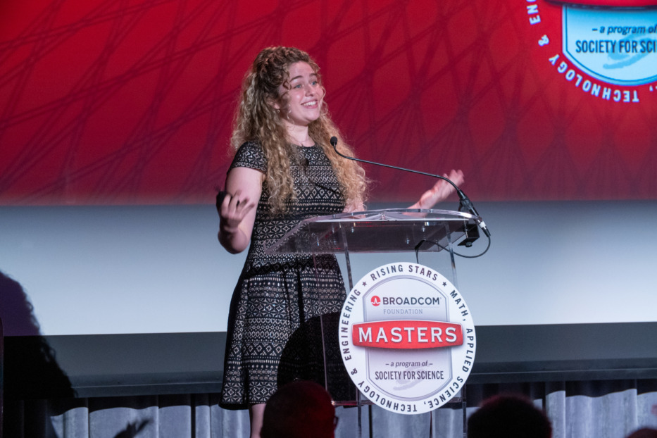 Broadcom Masters Alumna is speaking at a podium with a red back drop.