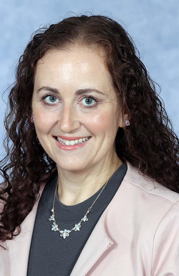 Woman with long brown curly hair smiling and wearing a pink blazer and necklace with a blue backdrop.