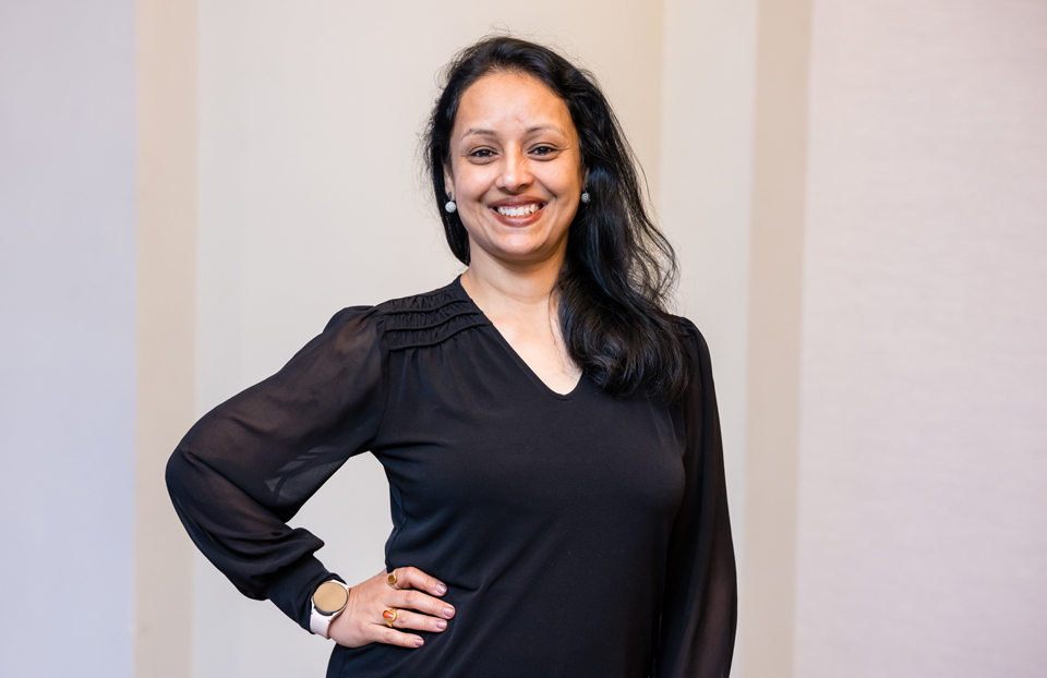 Woman with dark brown hair smiling at the camera wearing a black top and hand on her hip. 
