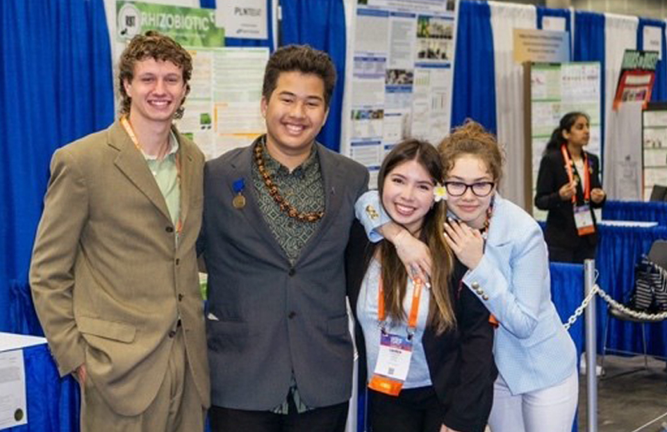 Two young men and two young girls are posing in front of science project boards in a group photo.
