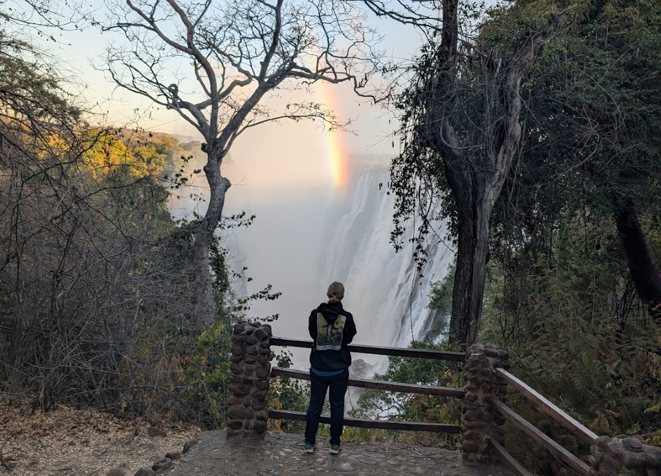 Leif standing on a bridge in Zambia, looking toward a waterfall and a rainbow.